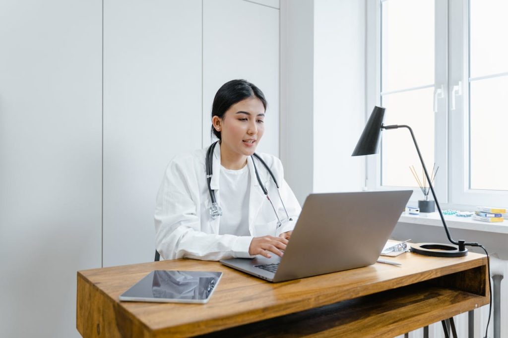 a-woman-in-white-coat-using-a-macbook-8376232 A female doctor in a white coat uses a laptop for an online consultation from her office.